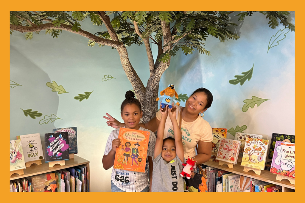 A mom, daughter and son smile with books and book-character stuffed animals in front of a faux tree in a bookstore.