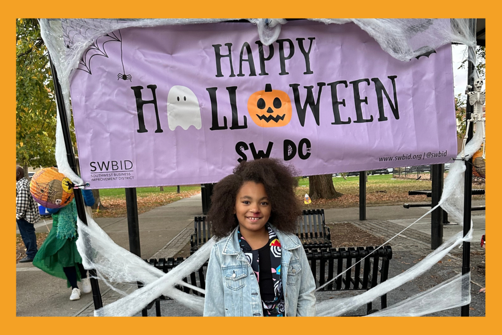 a girl in an eleven costume smiles with a faux bloody nose in front of a Happy Halloween SW DC sign.