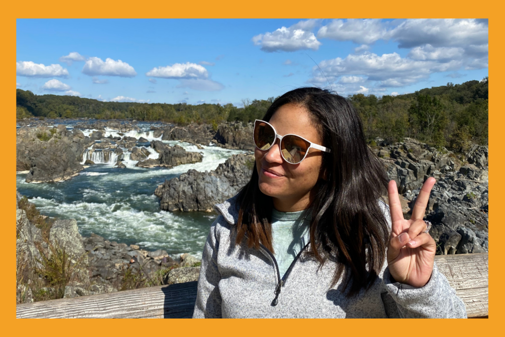 a woman holds a peace sign up in front of a beautiful waterfall at Great Falls Park.