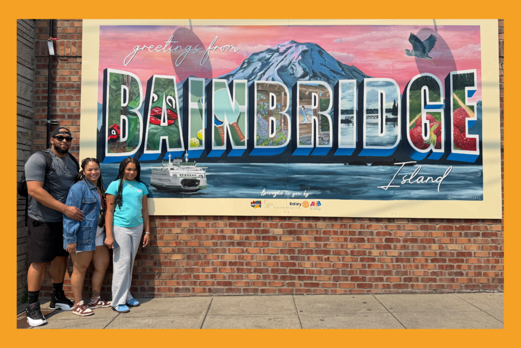 mom, dad, and daughter pose in front of the greetings from Bainbridge island mural.