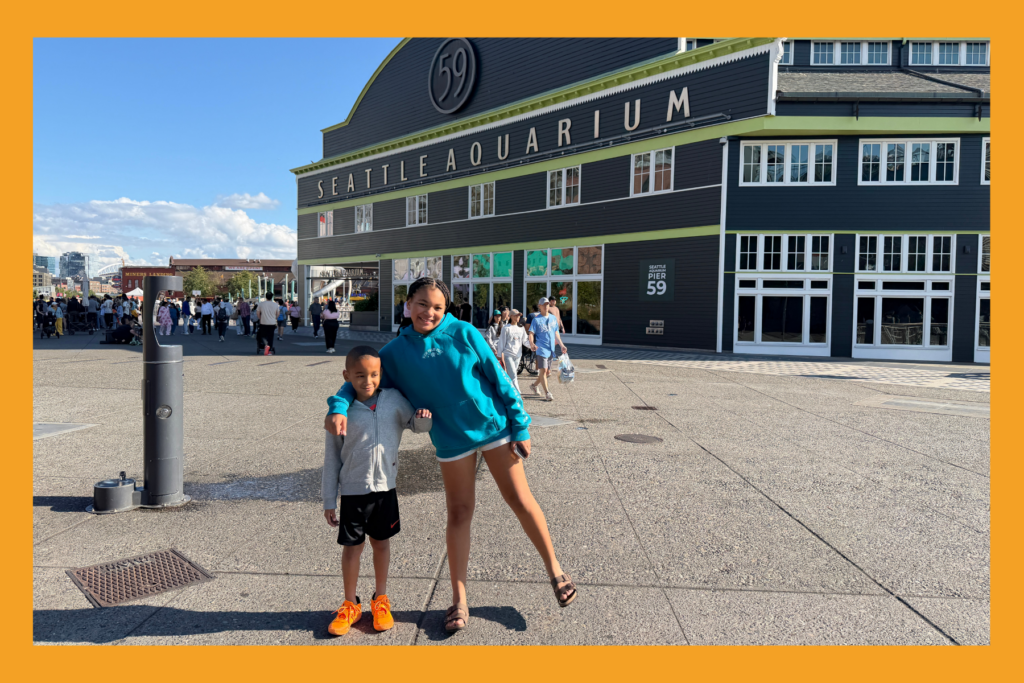 A sister places her arm around her brother in front of the Seattle Aquarium.
